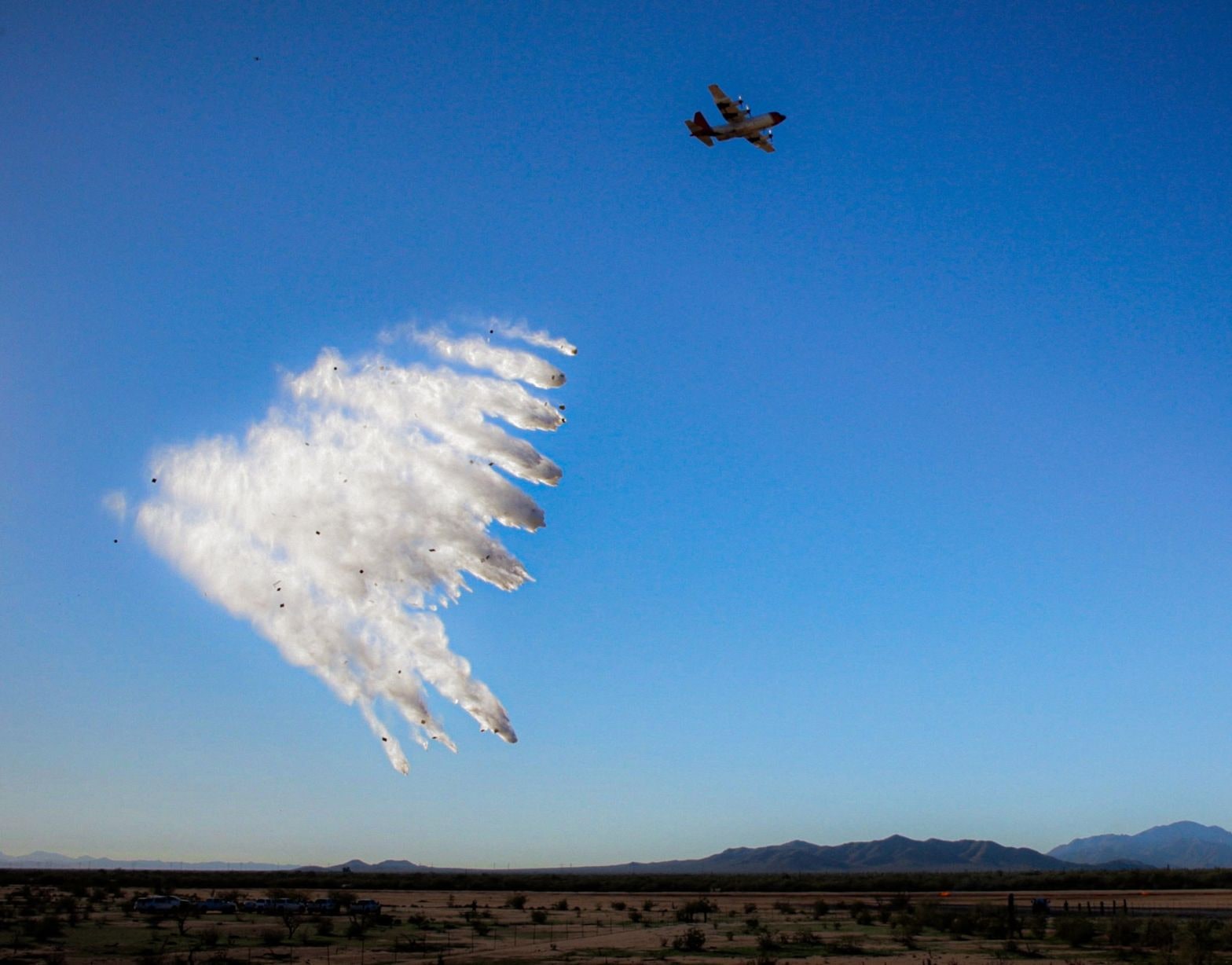 Full stick of PCADS containers rupturing into a retardant curtain below a C-130 over desert terrain