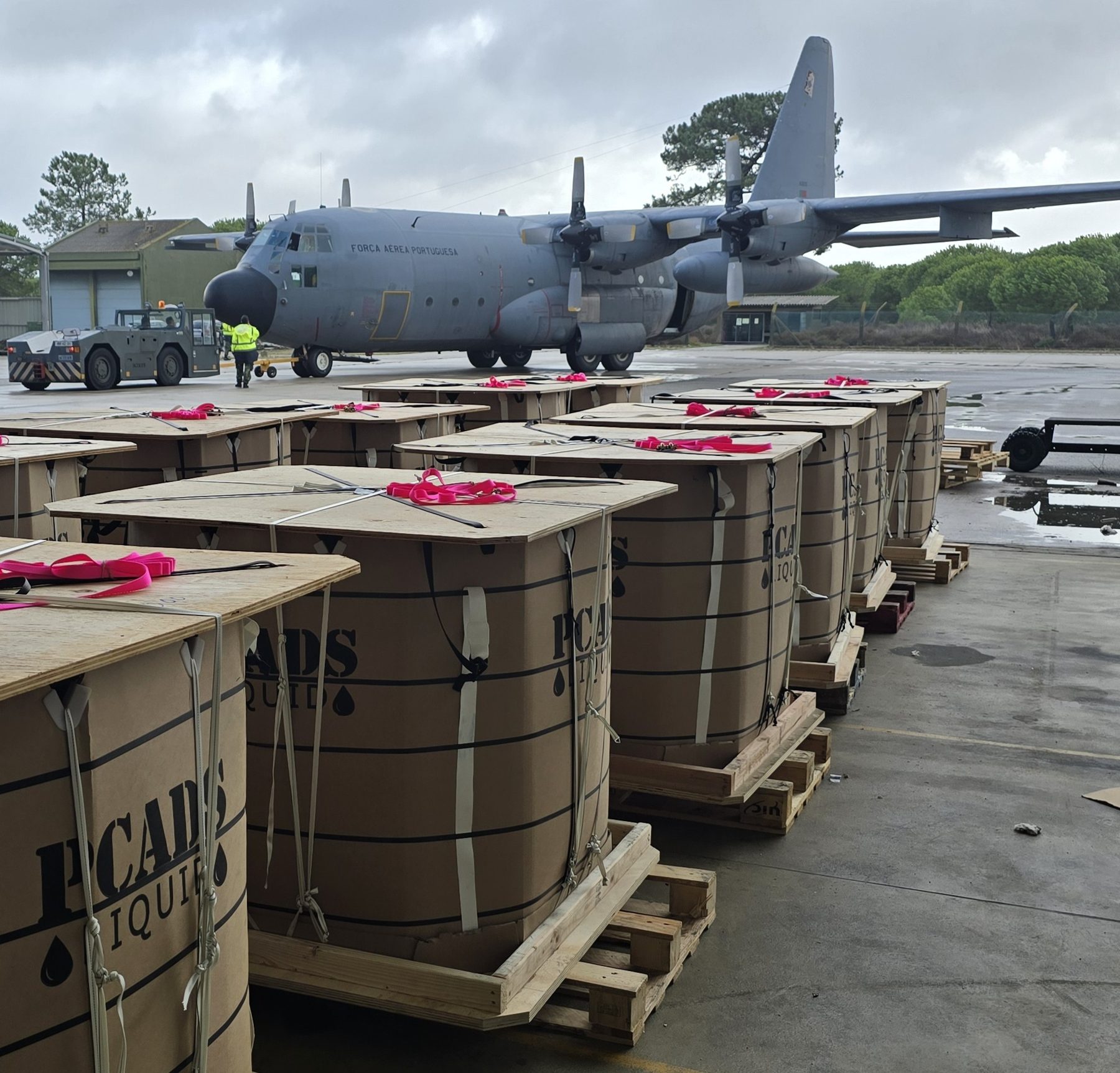 Rows of palletised PCADS Liquid containers staged on a wet flight line beside a Portuguese Air Force C-130