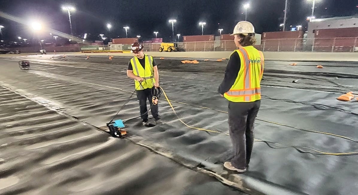 Crew wedge-welding containment liner seams at night under floodlights at a container terminal