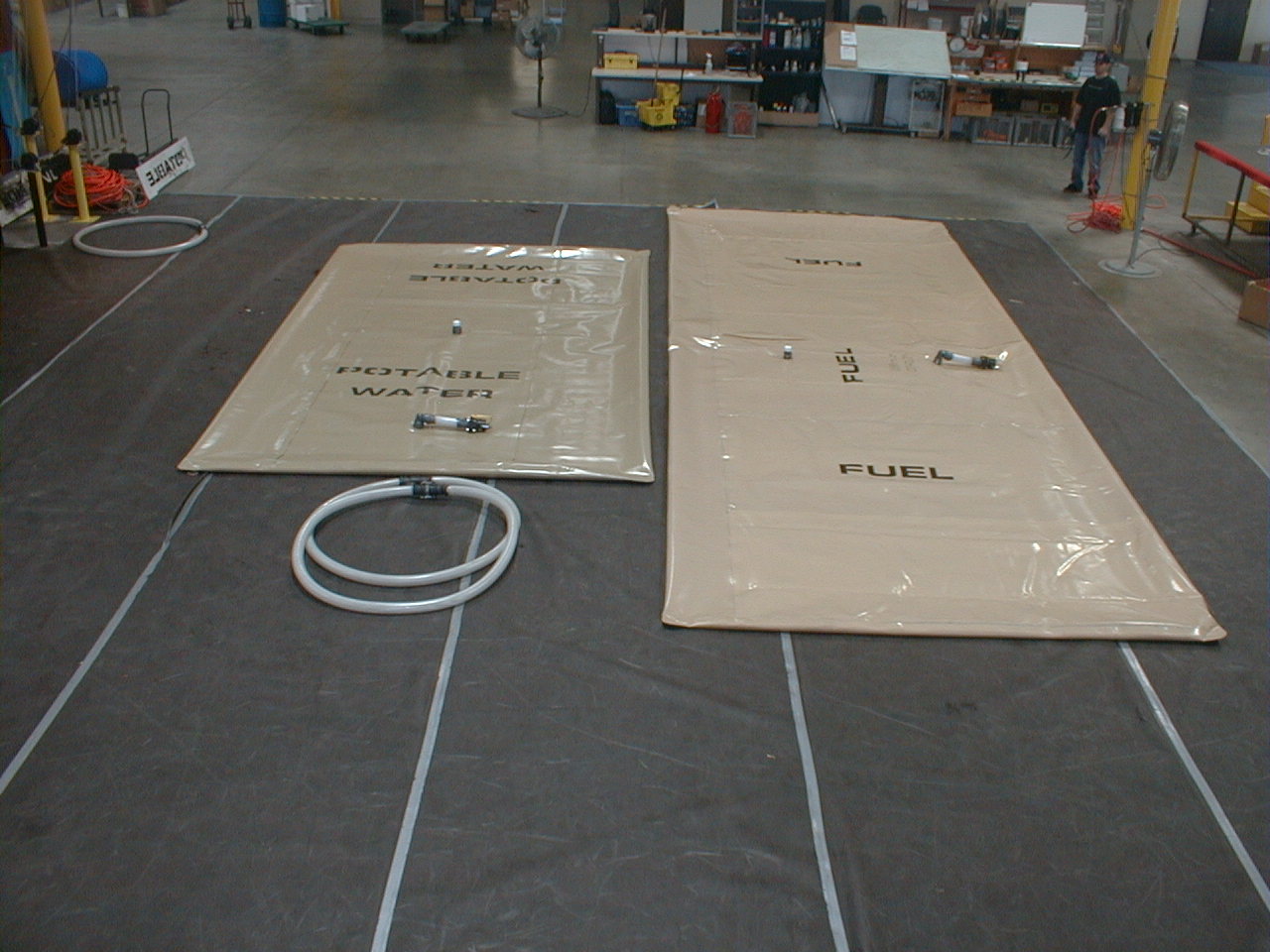 A fuel tank and a potable water tank laid out flat on the shop floor with hoses and fittings